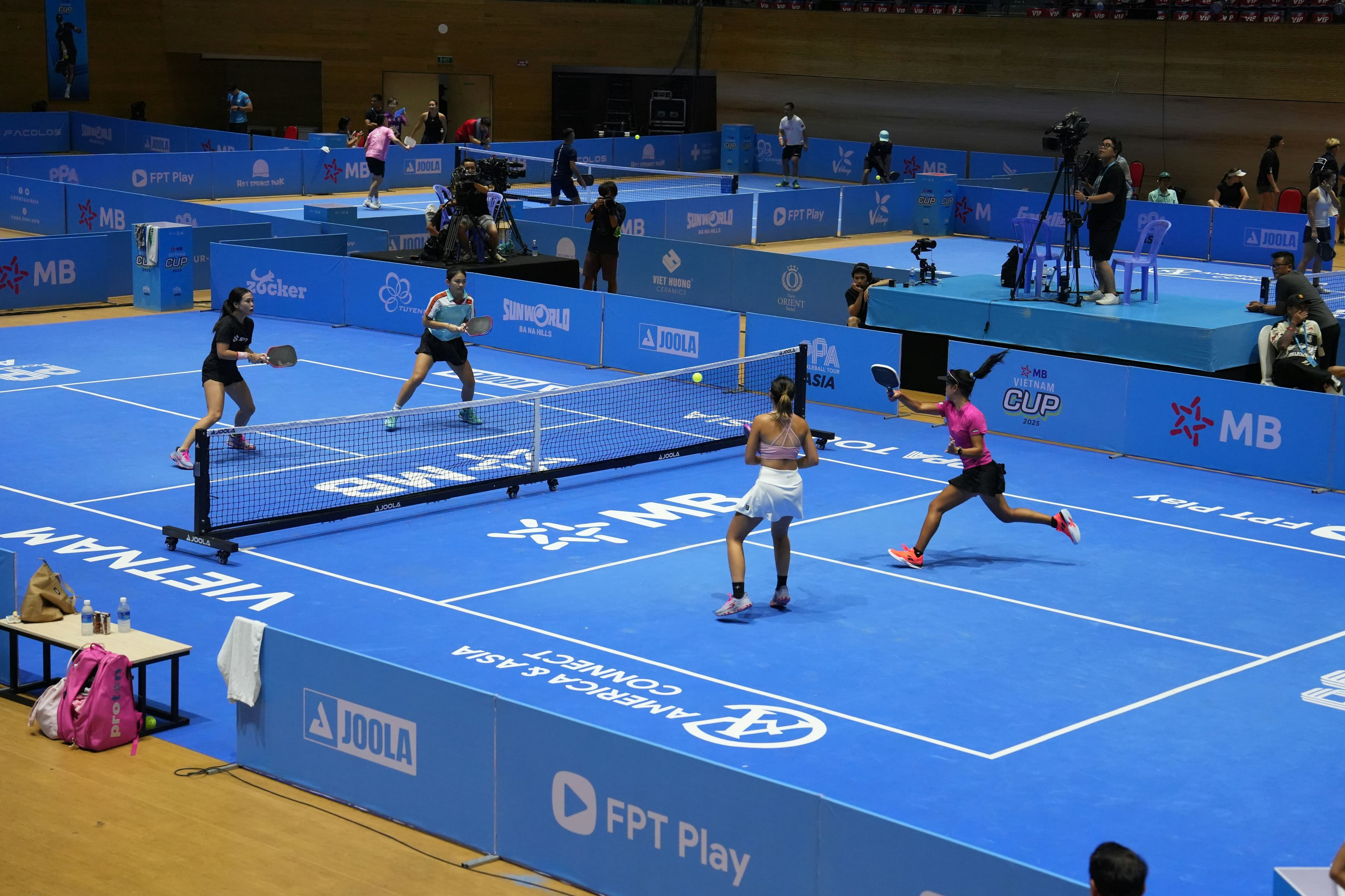 A refereed doubles match on a pro tournament court, with camera crews and more courts behind